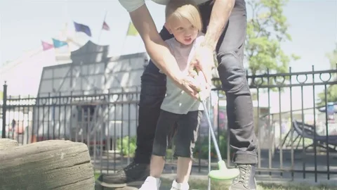 Loving father teaches his kid how to hit a golf ball at a putt putt golf course Stock Footage