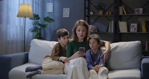 Loving mother using smartphone reading book to her children in the evening Stock Footage 264397920