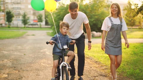 Loving parents are teaching son cute boy to ride bike in city park, child is Stock Footage 93134982