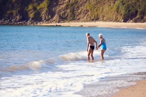 Loving Senior Couple Holding Hands As They Walk Into Sea For Swim Stock Photos