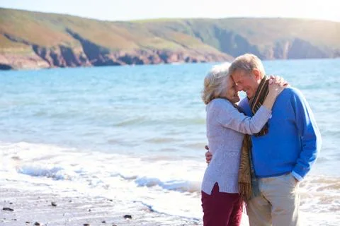 Loving Senior Couple Hugging As They Walk Along Shoreline Of Beach By Waves Stock Photos