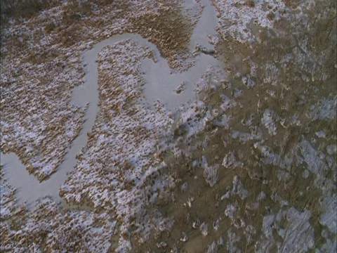 Low aerial over snow covered marsh land with frozen river meandering through. Stock Footage 86750545