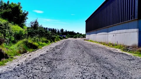 Low angle aerial dolly forward along gravel path by industrial building 库存影片 335684972
