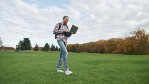 Low angle of concentrated young man walking on green lawn typing on laptop Stock Footage 256045444