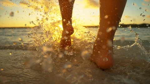 LOW ANGLE: Glassy drops of water splash around the girl's feet on sunny evening. Stock Footage 99558475