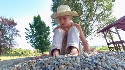 Low Angle Medium Static Shot of Girl in Straw Hat Burying Bare Feet in Pebbles 動画素材 346421672
