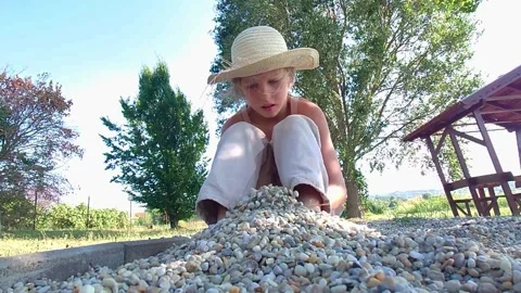 Low Angle Medium Static Shot of Young Girl in Straw Hat Playing with Pebbles in 動画素材 346421677