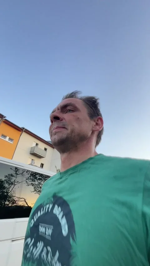 Low-angle shot of man in green t-shirt walking past apartment building at dusk Видео 344584052