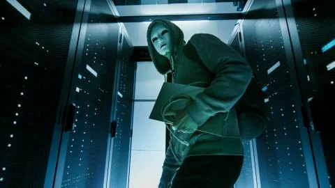 Low Angle Shot of a Masked Hacker in a Hoodie Sneaking Through Corporate Data Stock Photos