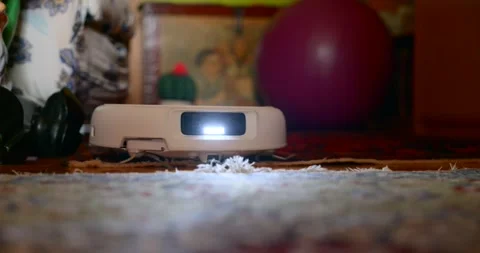 Low-angle shot of a robot vacuum passing under furniture, LED sensor glowing as Video stock 328533960