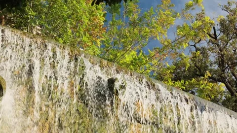 Low angle view of an artificial waterfall flowing down a stone wall, with lush Stock Footage 347025713