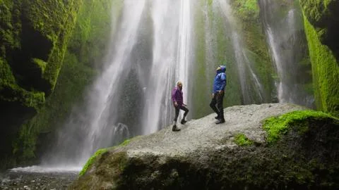 Low angle view of couple standing on mossy boulder admiring waterfall in remote 库存照片
