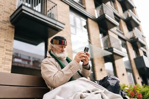 Low-angle view of elderly homeless man sitting on urban bench by luxury Stock Photos
