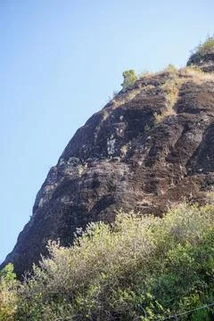 Low angle view of a giant black rock cliff overgrown with wild vegetation Stock Photos
