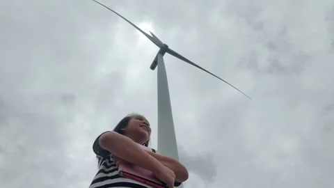 Low angle view of happy Asian girl looking up at giant rotating wind turbine Video stock 343197231