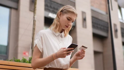 Low-angle view of smiling female tourist holding bank card, entering cvv code or Stock Footage 228473370