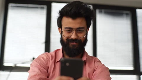 Low-angle view of smiling Indian hipster male in glasses using smartphone Stock Footage 172502360