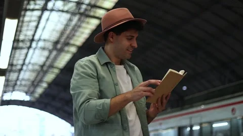 Low-angle view of traveler with hat reading book inside train station, 4k vid Stock Footage 298562194