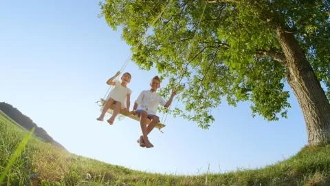 LOW ANGLE VIEW Two happy kids swinging on a swing under a big tree on a warm day Stock Photos