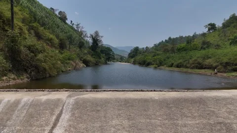 Low drone flight upstream over dry season spillway with mountain backdrop 動画素材 336973600