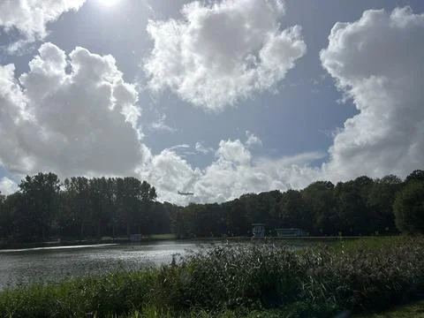 Low flying passenger plane above tree lined lake under cumulus clouds, reeds Stock Photos