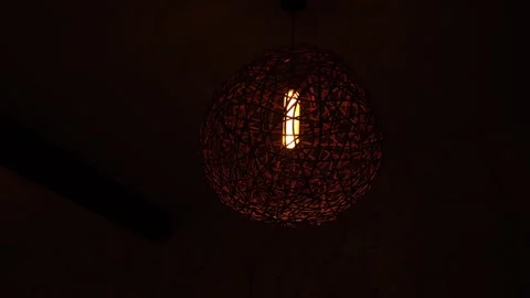 A low-light close-up of a woven spherical pendant lamp with a warm glowing bulb Stock Footage 332974667