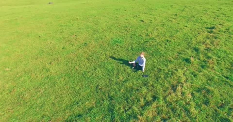 Low orbital flight around man on green grass with notebook pad at yellow rural Stock Footage 285477584