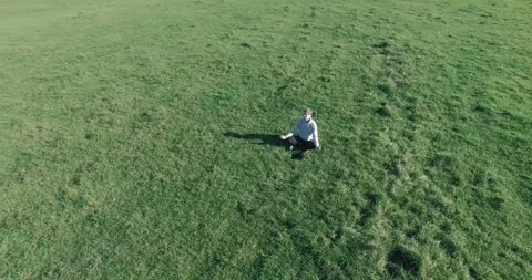 Low orbital flight around man on green grass with notebook pad at yellow rural Stock Footage 291782643