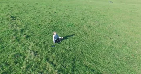 Low orbital flight around man on green grass with notebook pad at yellow rural Stock Footage 291782716