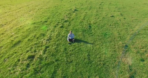 Low orbital flight around man on green grass with notebook pad at yellow rural Stock Footage 294421791