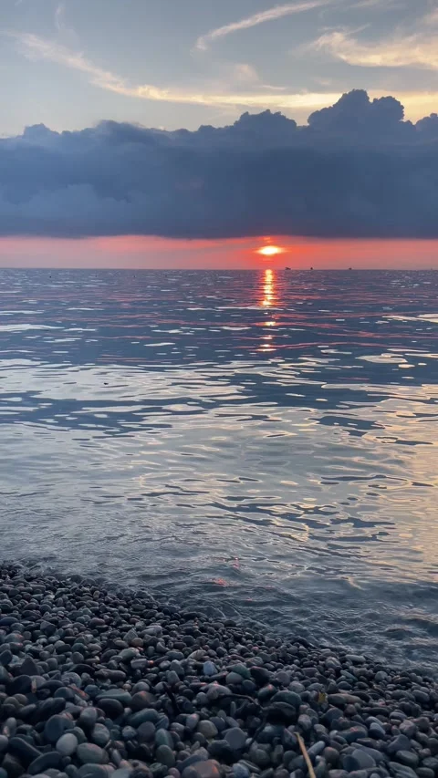 Low sun sets beyond a calm sea viewed from a pebble beach, while vivid red .. Stock Footage 341257405