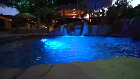 Low view across a glowing blue pool toward a rock waterfall Stock Footage 345539644