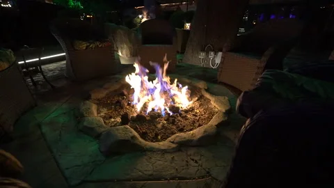 Low view of a patio fire pit with flames at night beside wicker chairs Stock Footage 345540801