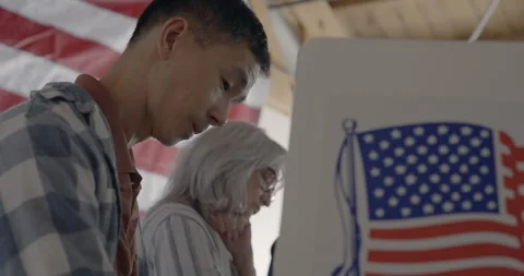 Low viewpoint, looking up at Asian man voting at booth, US flag behind Video stock 275202244