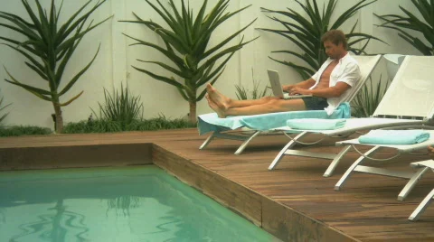 LS PAN OF A MAN WORKING ON A LAPTOP AND THEN DIPPING HIS FEET IN A SWIMMING POOL 스톡 동영상 543618