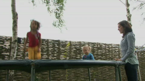 LS PAN OF A MOTHER WATCHING HER DAUGHTERS JUMPING ON A TRAMPOLINE Stock Footage