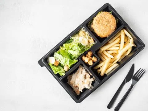 Lunch box with caesar salad, burger and fries in black plastic package Stock Photos