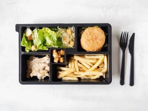 Lunch box with caesar salad, burger and fries in black plastic package Stock Photos