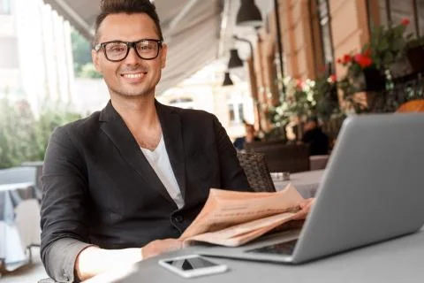 Lunch time. Young man in suit and glasses sitting at cafe on city street with Stock Photos