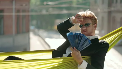 Ma in suit using waving fan in hammock during hot day Stock Footage 164036808