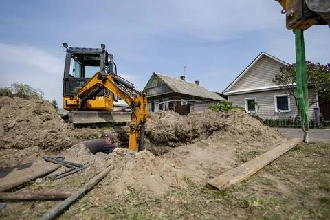 A machine is digging a deep hole in the ground next to two houses. A worker i Stock Photos