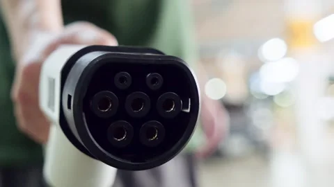 Macro shot of an EV charging plug held toward the lens, with blurred indoor Stock Footage 314770744