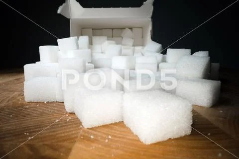 Macro shot of sugar cubes next to carton box on wooden table. Unhealthy food Stock Photos