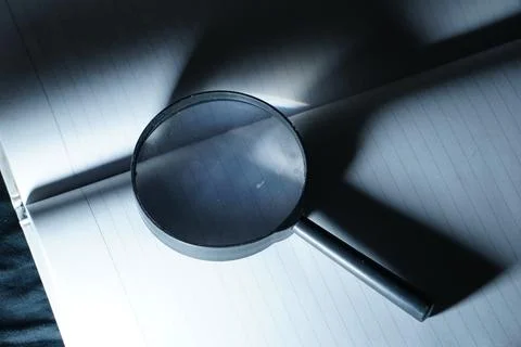 A magnifying glass is placed on an open notebook under a cool blue spotlight, cr Stock Photos