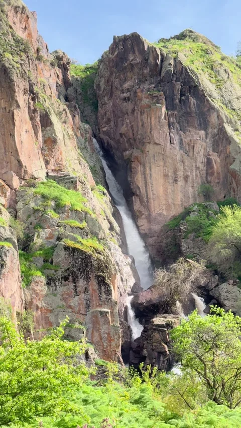 A majestic waterfall flows down a rugged cliff surrounded by lush greenery and Stock Footage 276426937
