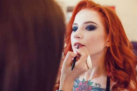 Make-up artist does makeup to the red-haired girl with tattoo in studio Stock Photos