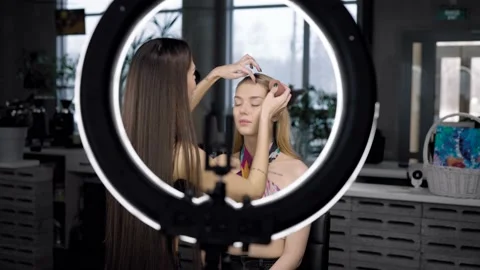 Make-up artist makes a makeup for a young blonde girl, illuminates with a ring Stock Footage 235217573