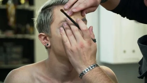 Makeup artist making face art for a young blonde man in beauty studio Stock Footage 76578873