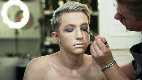 Makeup artist making face art for a young blonde man in the makeup studio Stock Footage 76579665