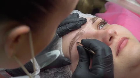 Makeup artist plucks eyebrows and doing eyebrow permanent makeup correction in a Stock Footage 262148120